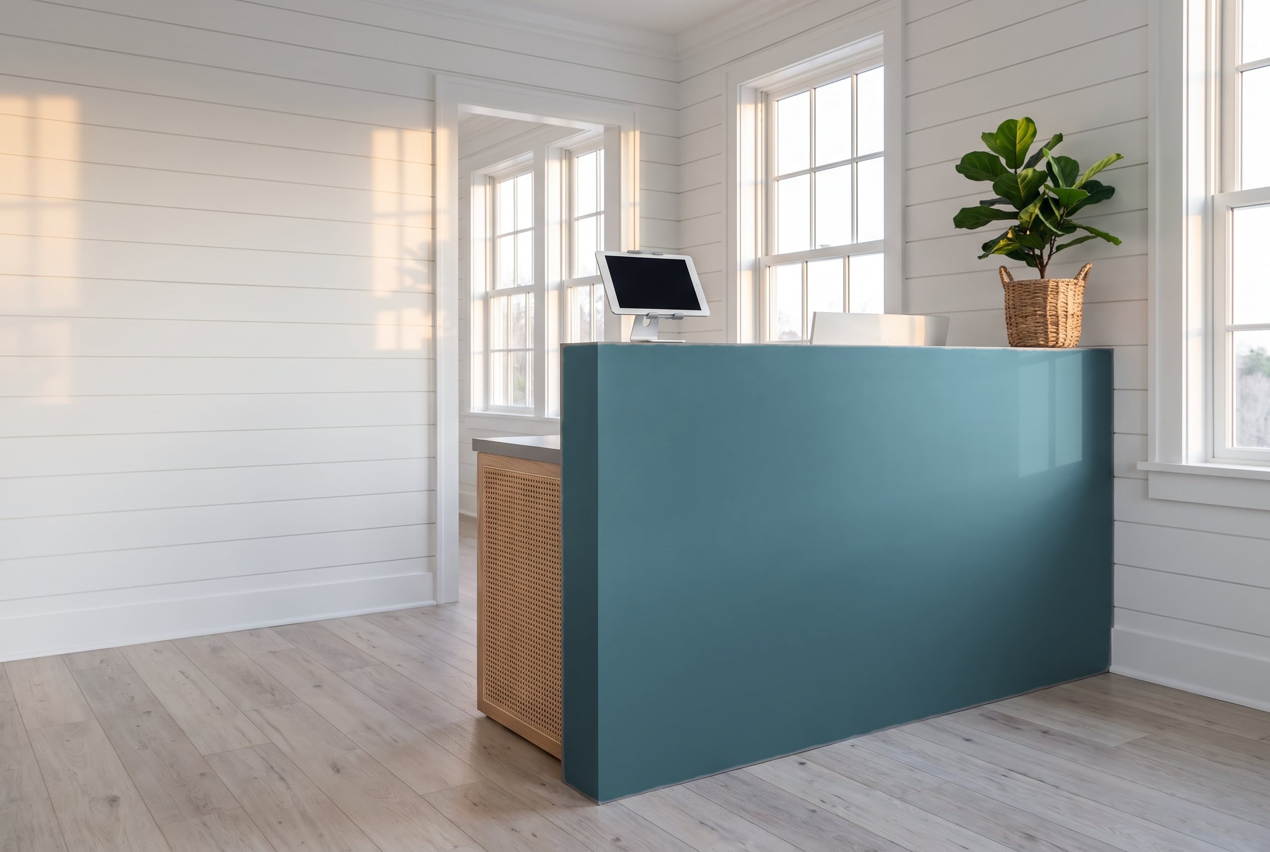 An empty reception desk with a tablet on a stand and a potted plant in a bright room with white plank walls.