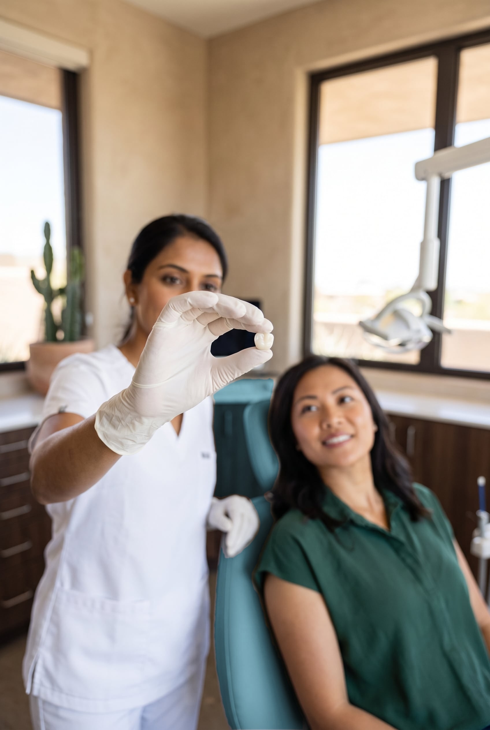 A gloved dentist holds up a small white tooth crown while a smiling woman watches from the dental chair.