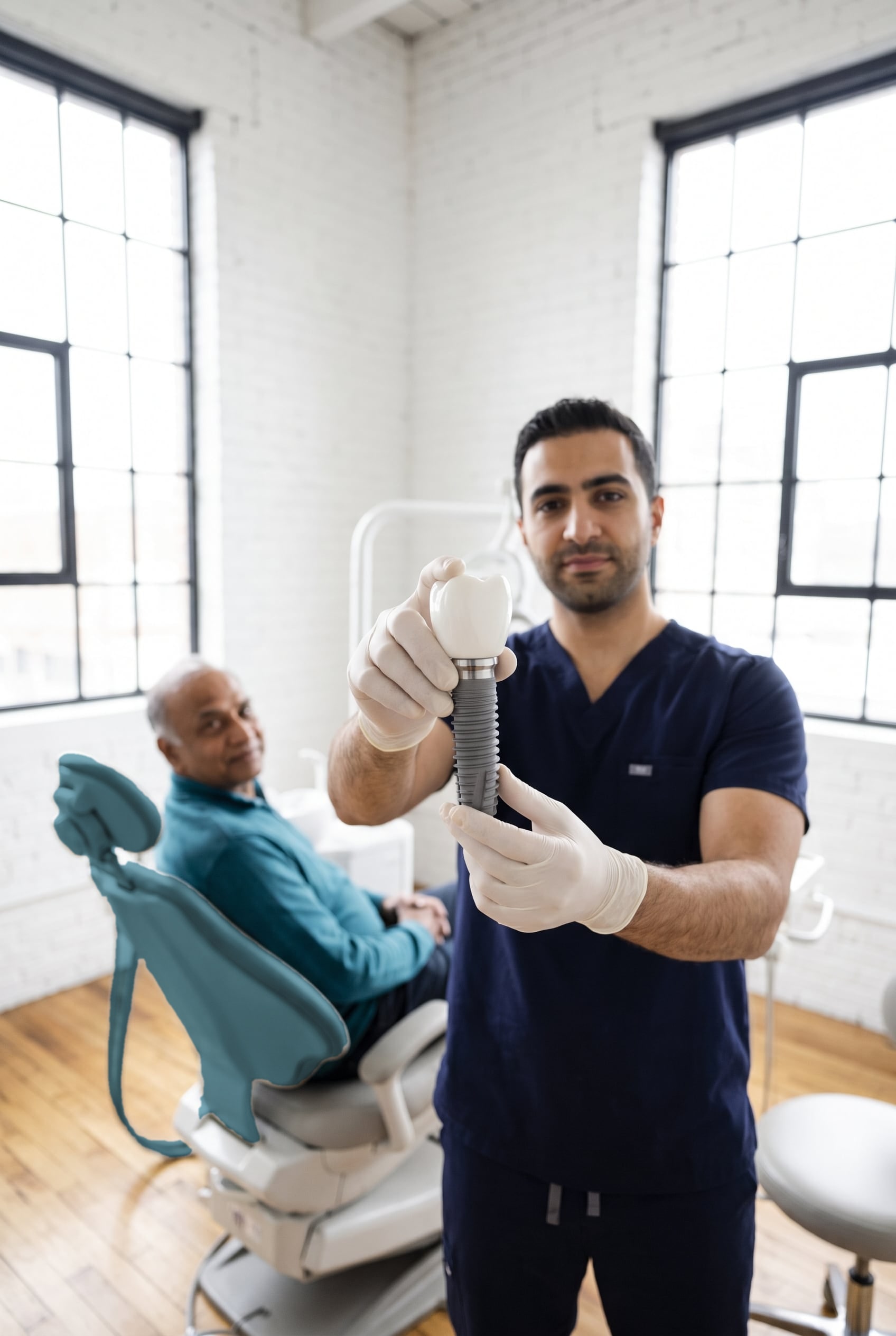 A gloved dentist holds up a large implant model while an older man waits in the dental chair behind him.