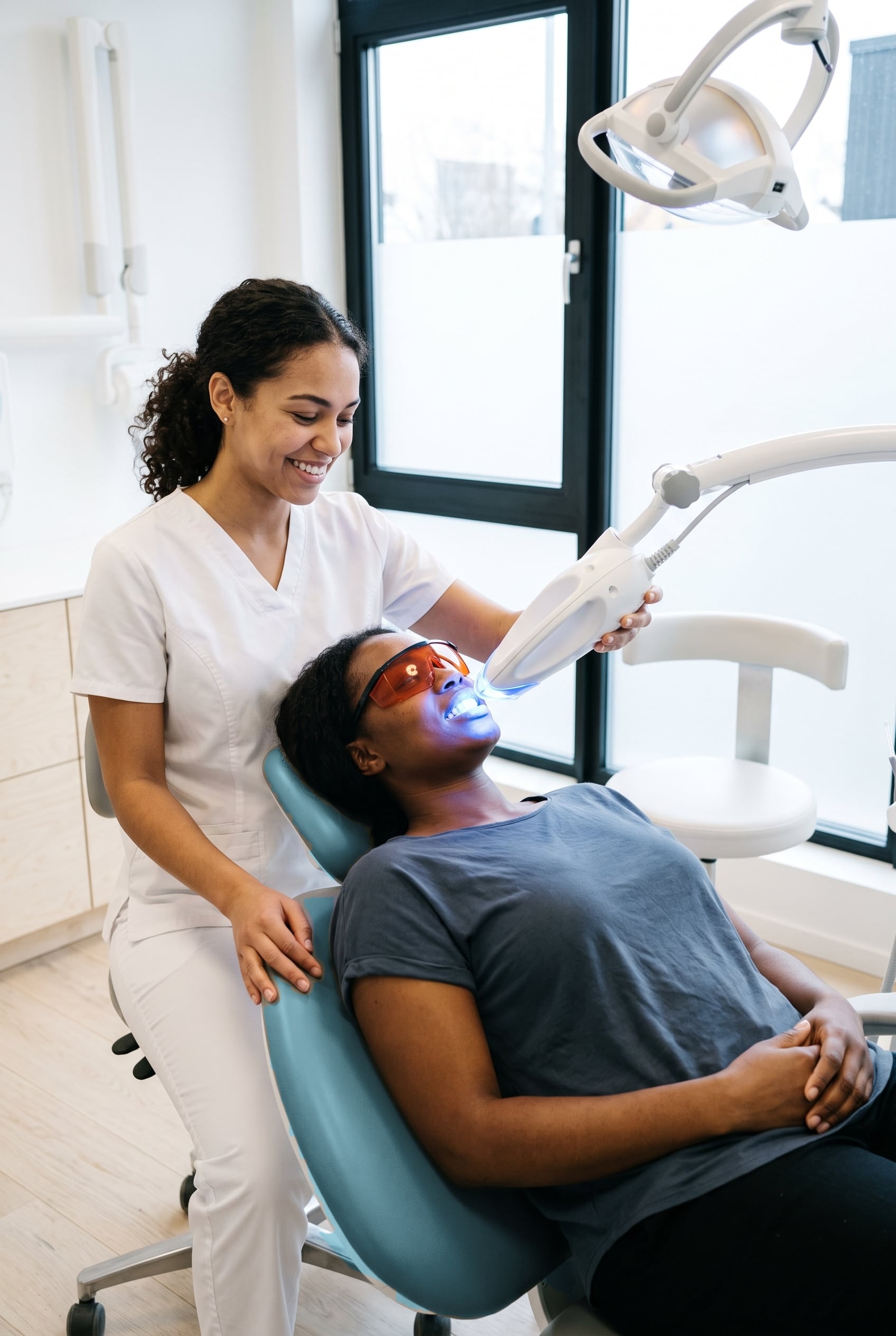 A woman in protective glasses reclines under a blue whitening light while a smiling dental assistant sits behind her.
